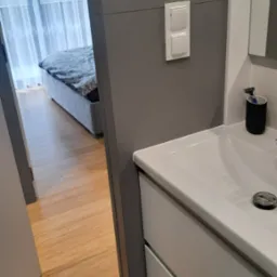 Nowoczesna łazienka z widokiem na sypialnię przez otwarte drzwi, szara ściana z panelem sterowania i włącznikami, biała umywalka z czarnym pojemnikiem na przybory do golenia, drewniana podłoga...