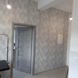 Ściana z tapetą w geometryczny wzór, szara drewniana ościeżnica z uchylonymi drzwiami, podłoga wyłożona jasnymi płytkami w odcieniach beżu.