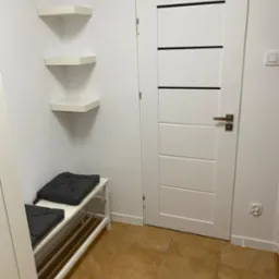 Przedpokój po remoncie: białe ściany, brązowe płytki podłogowe, białe drzwi z poziomymi czarnymi paskami, narożne półki i biała ławeczka z szarymi poduszkami.