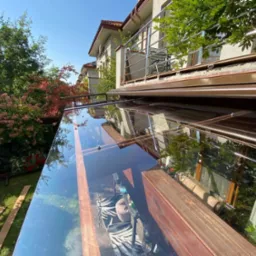 Szklana konstrukcja z drewnianymi elementami, prawdopodobnie zadaszenie tarasu, z widokiem na ogród i budynek mieszkalny w tle.