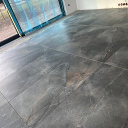 Stivbud - Ułożone, ciemnoszare płytki podłogowe z marmurkowym wzorem w nowym pomieszczeniu z oknem zabezpieczonym folią ochronną i widocznymi gniazdkami elektrycznymi.