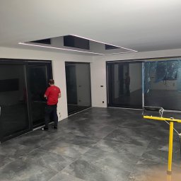 Stivbud - Wnętrze w trakcie prac wykończeniowych: pracownik w czerwonej koszulce reguluje przesuwne drzwi balkonowe na tle szarej, marmurkowej podłogi i sufitu z podświetleniem LED.