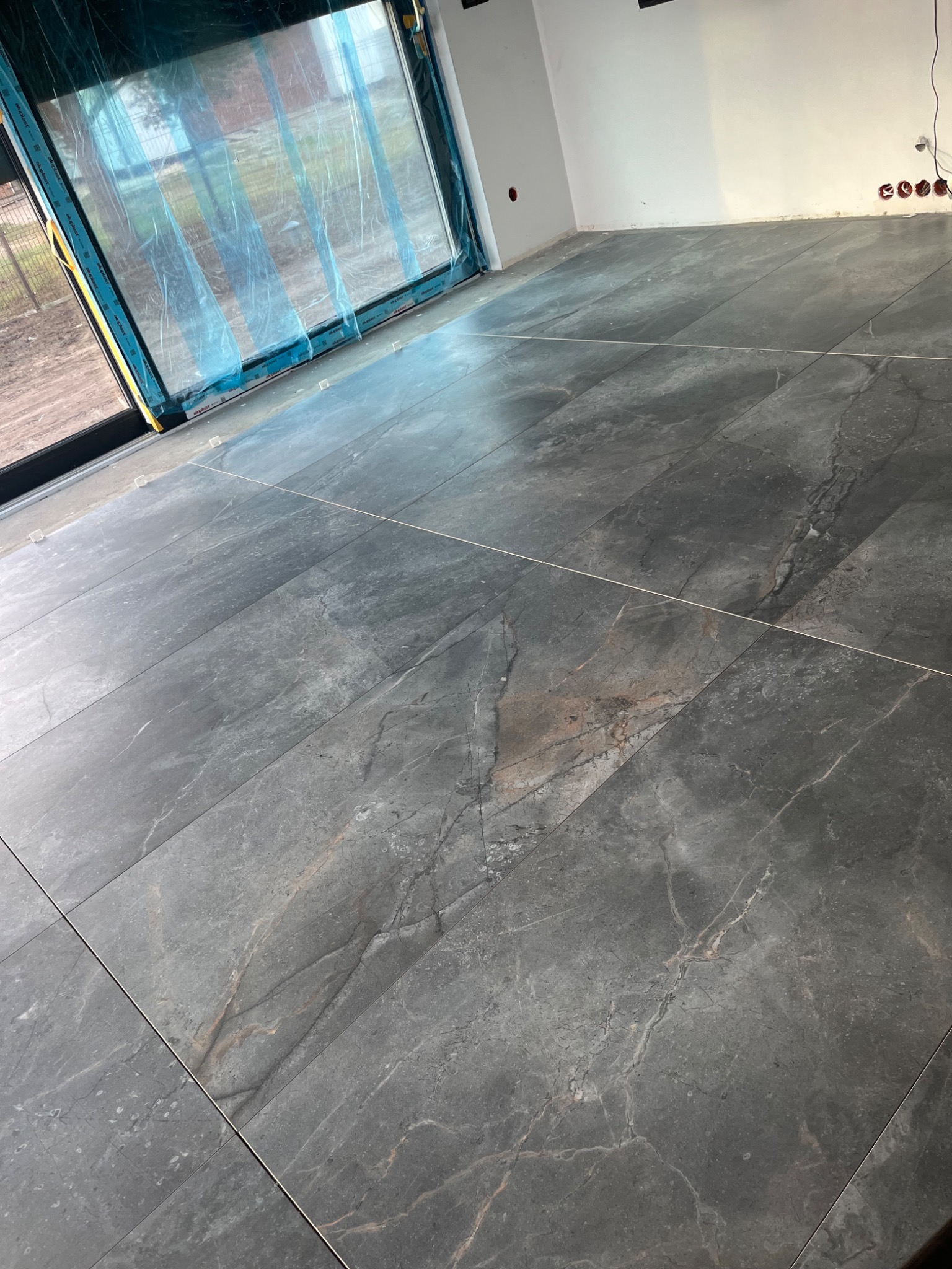 Ułożone, ciemnoszare płytki podłogowe z marmurkowym wzorem w nowym pomieszczeniu z oknem zabezpieczonym folią ochronną i widocznymi gniazdkami elektrycznymi.