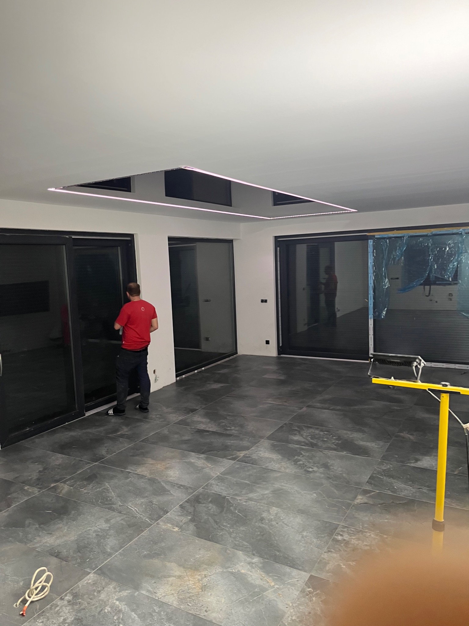 Wnętrze w trakcie prac wykończeniowych: pracownik w czerwonej koszulce reguluje przesuwne drzwi balkonowe na tle szarej, marmurkowej podłogi i sufitu z podświetleniem LED.