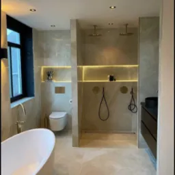 Nowoczesna łazienka z wolnostojącą wanną, kabiną prysznicową typu walk-in z dwoma deszczownicami i podświetlanymi niszami, wykończona w beżowych płytkach imitujących marmur.