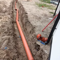 Pomarańczowe rury kanalizacyjne ułożone w wykopie ziemnym, widoczne złącza i kształtki, fragment ściany budynku z rurą spustową w tle.