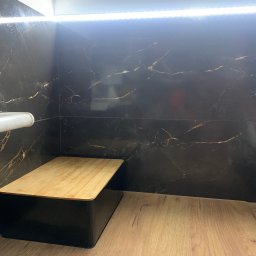 bTb constructions - Fragment wykończonej łazienki z czarnymi płytkami imitującymi marmur, podświetleniem LED i drewnianym podestem z napisem 'Villa Ambition'.