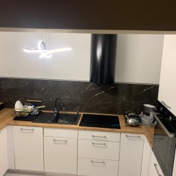 bTb constructions - Nowoczesna kuchnia z białymi szafkami, drewnianym blatem, czarną armaturą i marmurkową ścianą nad blatem, oświetlona designerską lampą wiszącą.