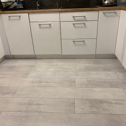 bTb constructions - Ułożone jasnoszare panele podłogowe przypominające drewno w kuchni z białymi szafkami i drewnianym blatem, widok z niskiej perspektywy.