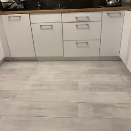 Ułożone jasnoszare panele podłogowe przypominające drewno w kuchni z białymi szafkami i drewnianym blatem, widok z niskiej perspektywy.