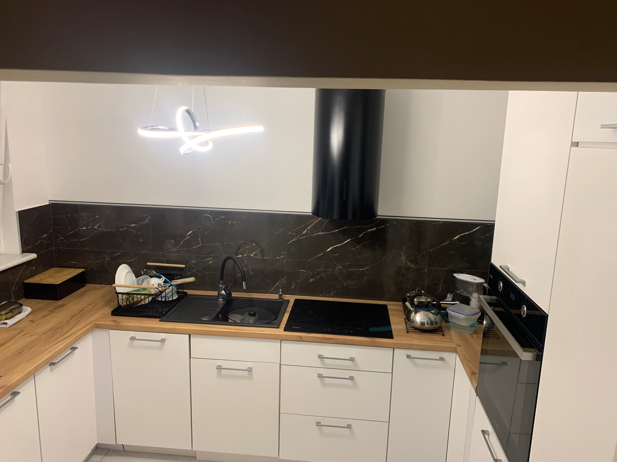 Nowoczesna kuchnia z białymi szafkami, drewnianym blatem, czarną armaturą i marmurkową ścianą nad blatem, oświetlona designerską lampą wiszącą.