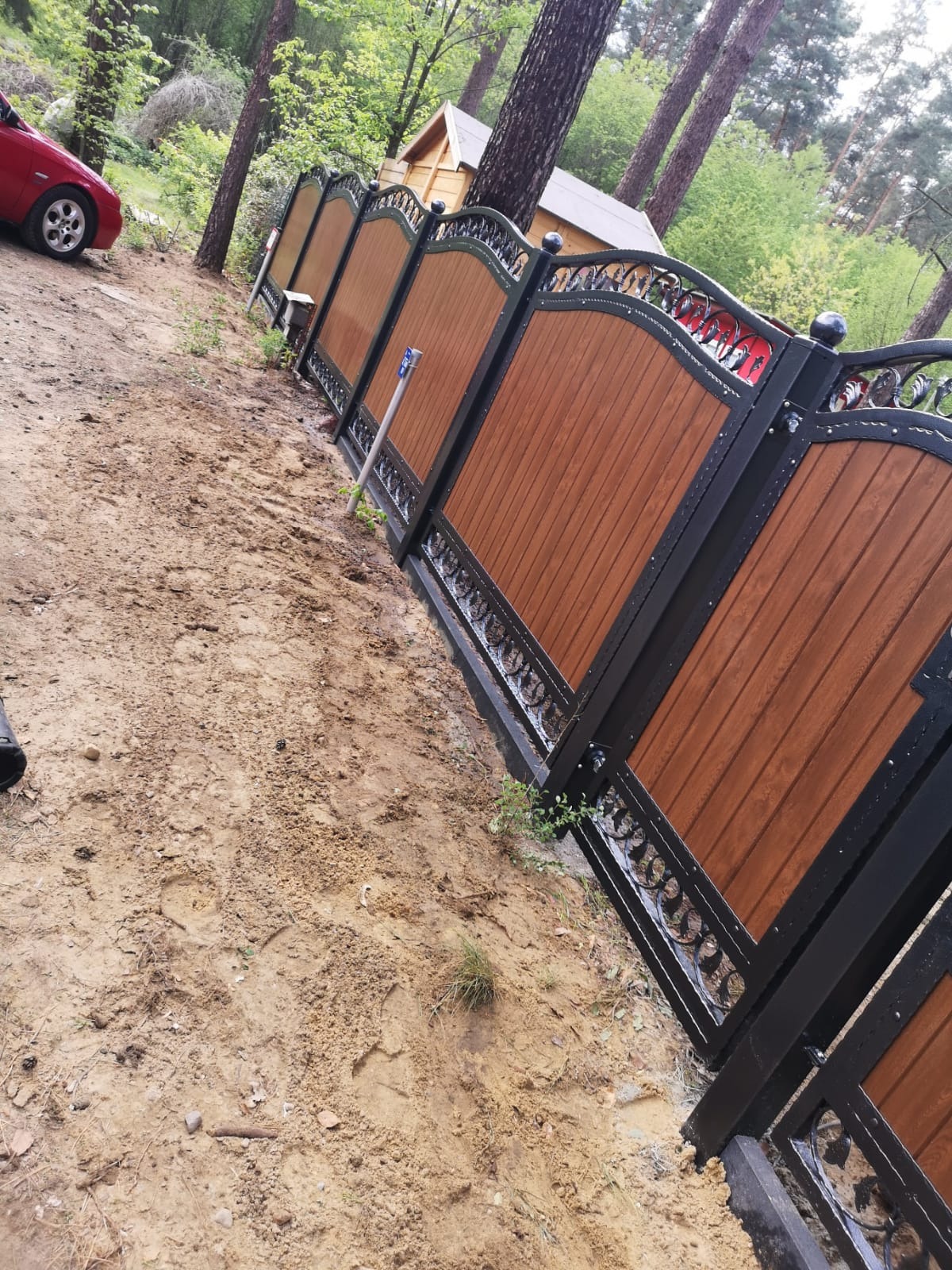 Nowoczesna brama wjazdowa z drewnianymi panelami i ozdobnymi elementami kutymi, widoczna na tle leśnej działki z czerwonym samochodem w oddali.