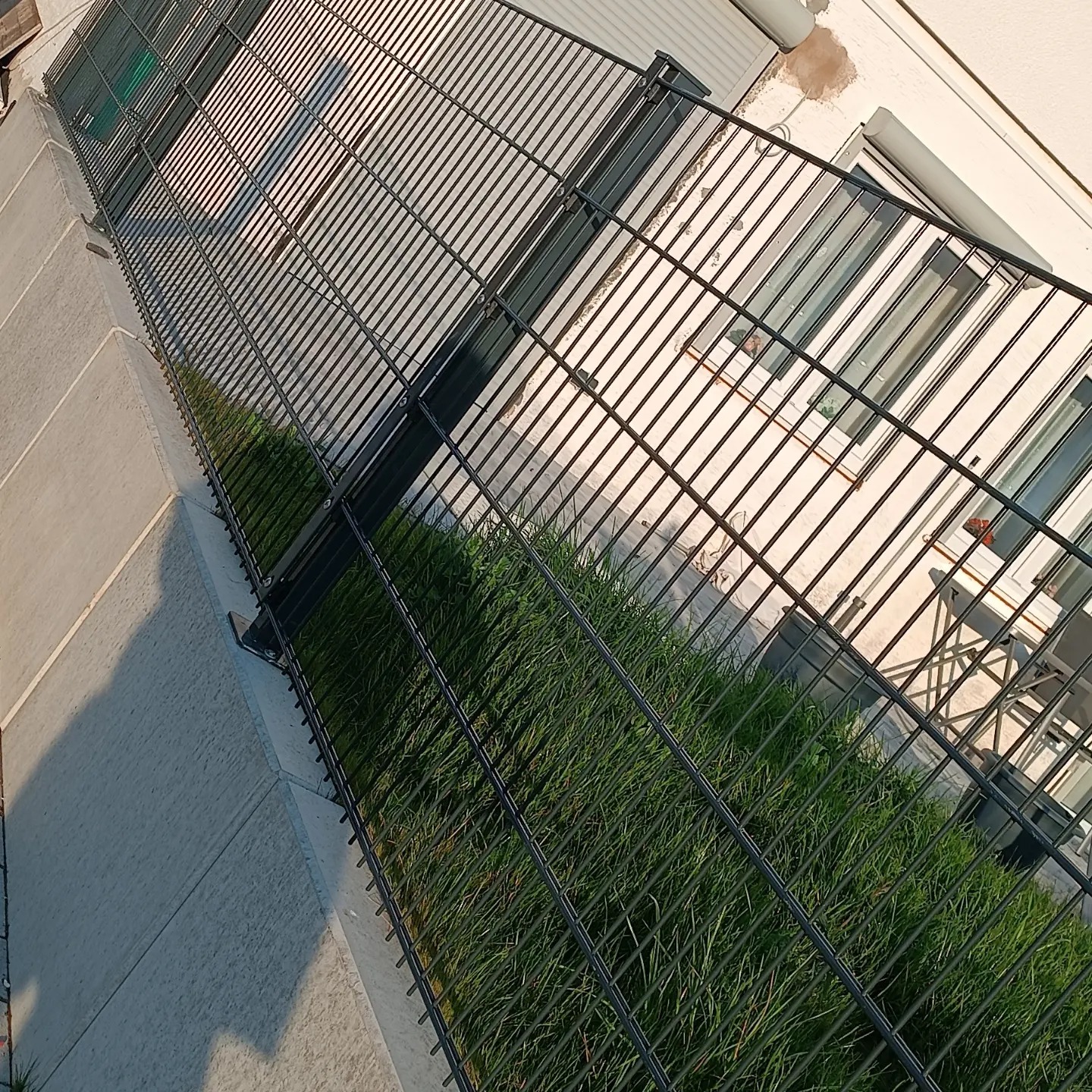 Nowoczesne, czarne ogrodzenie z paneli siatkowych, widok z góry, zza ogrodzenia widoczny trawnik, fragment budynku z oknami i meblami ogrodowymi.