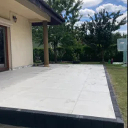 Nowoczesny taras z jasnych płyt, obrzeża z ciemnej kostki, widok z poziomu trawnika, w tle dom z kolumną i zieleń ogrodu.