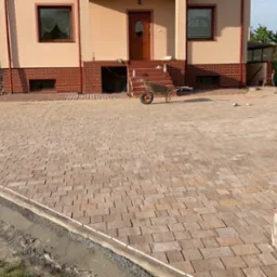 Ułożona kostka brukowa w kolorze beżowo-brązowym przed domem jednorodzinnym; na pierwszym planie stos kostki, w tle pracownik przy maszynie do cięcia kostki.