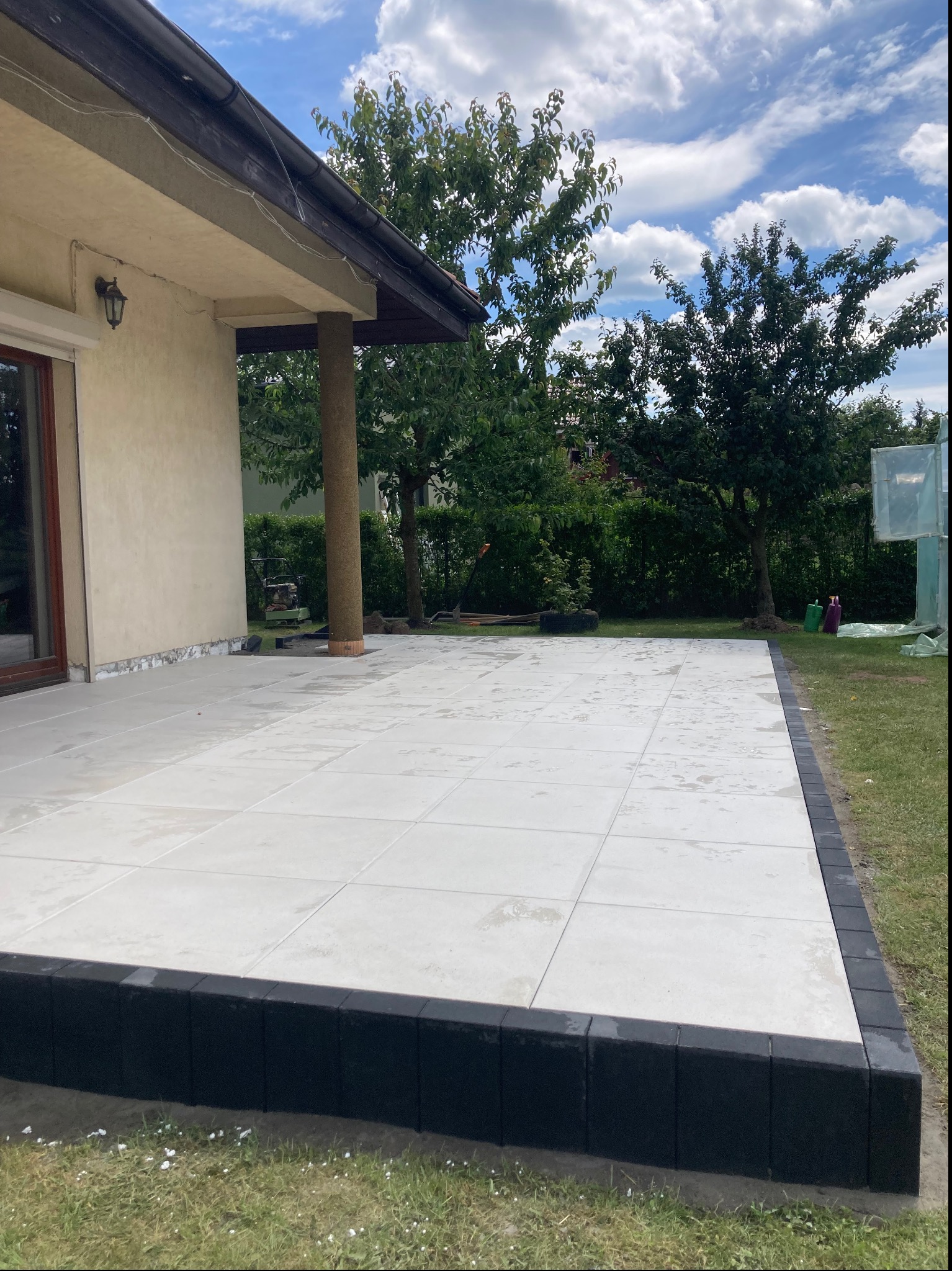 Nowoczesny taras z jasnych płyt, obrzeża z ciemnej kostki, widok z poziomu trawnika, w tle dom z kolumną i zieleń ogrodu.