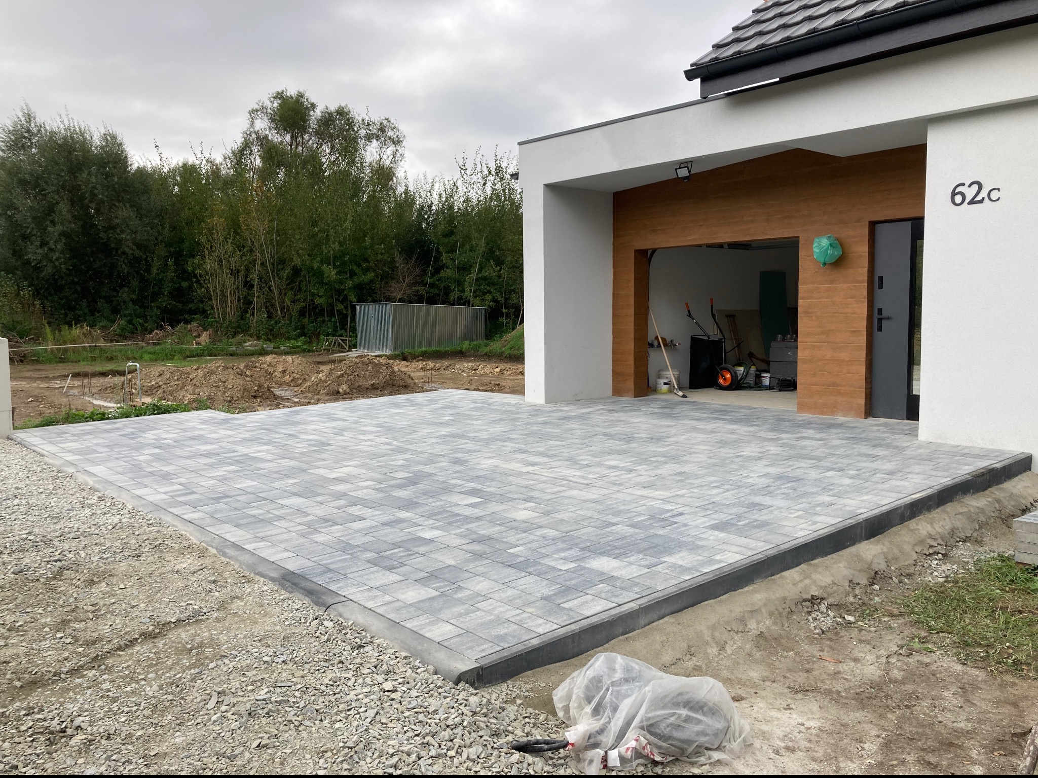 Nowo ułożona, szara kostka brukowa przed nowoczesnym domem z numerem 62c, widoczne przygotowane podłoże i narzędzia w folii.
