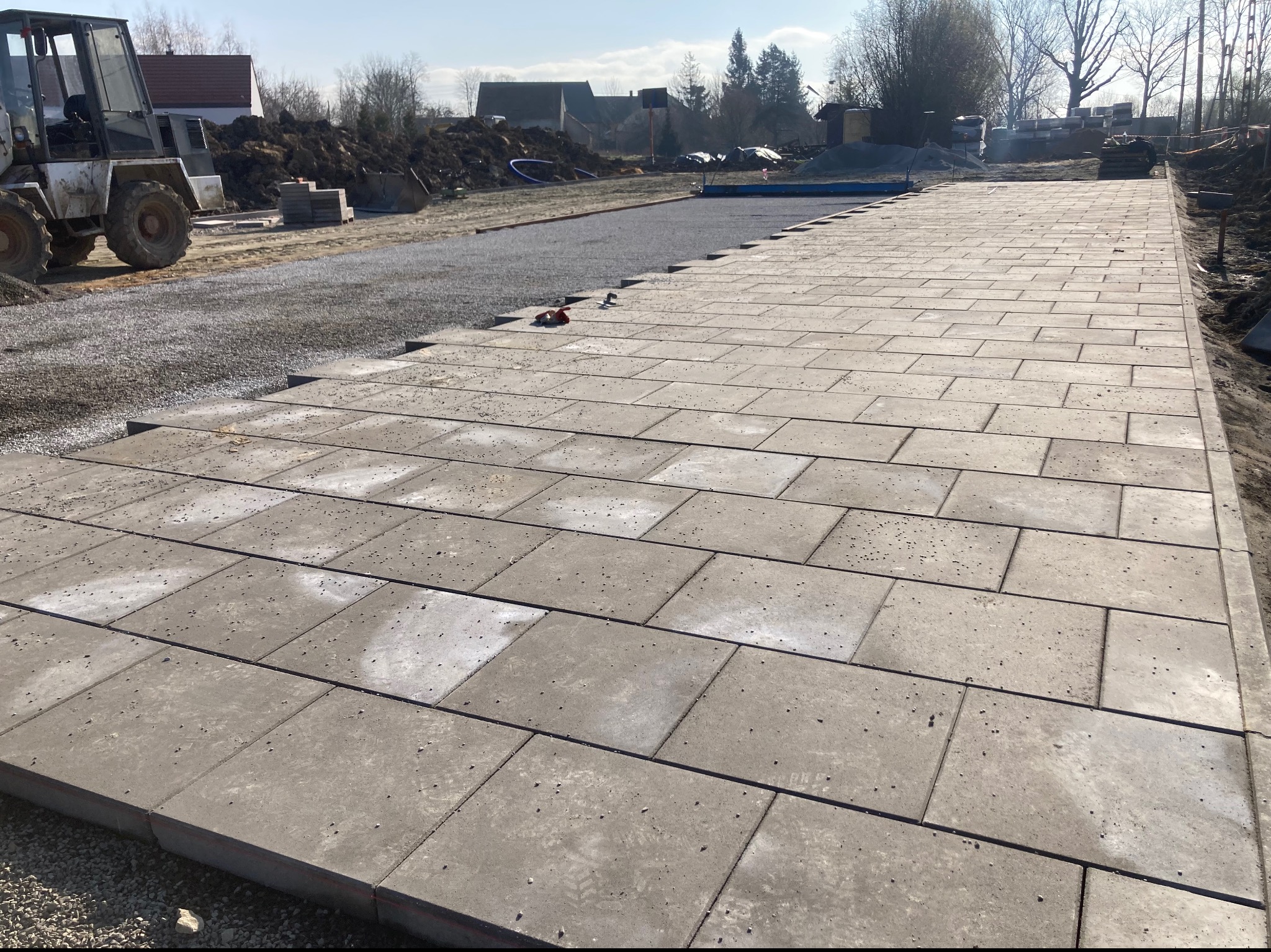 Prostokątna, szara kostka brukowa ułożona na podsypce z kruszywa, widoczna maszyna budowlana i przygotowany teren w tle.