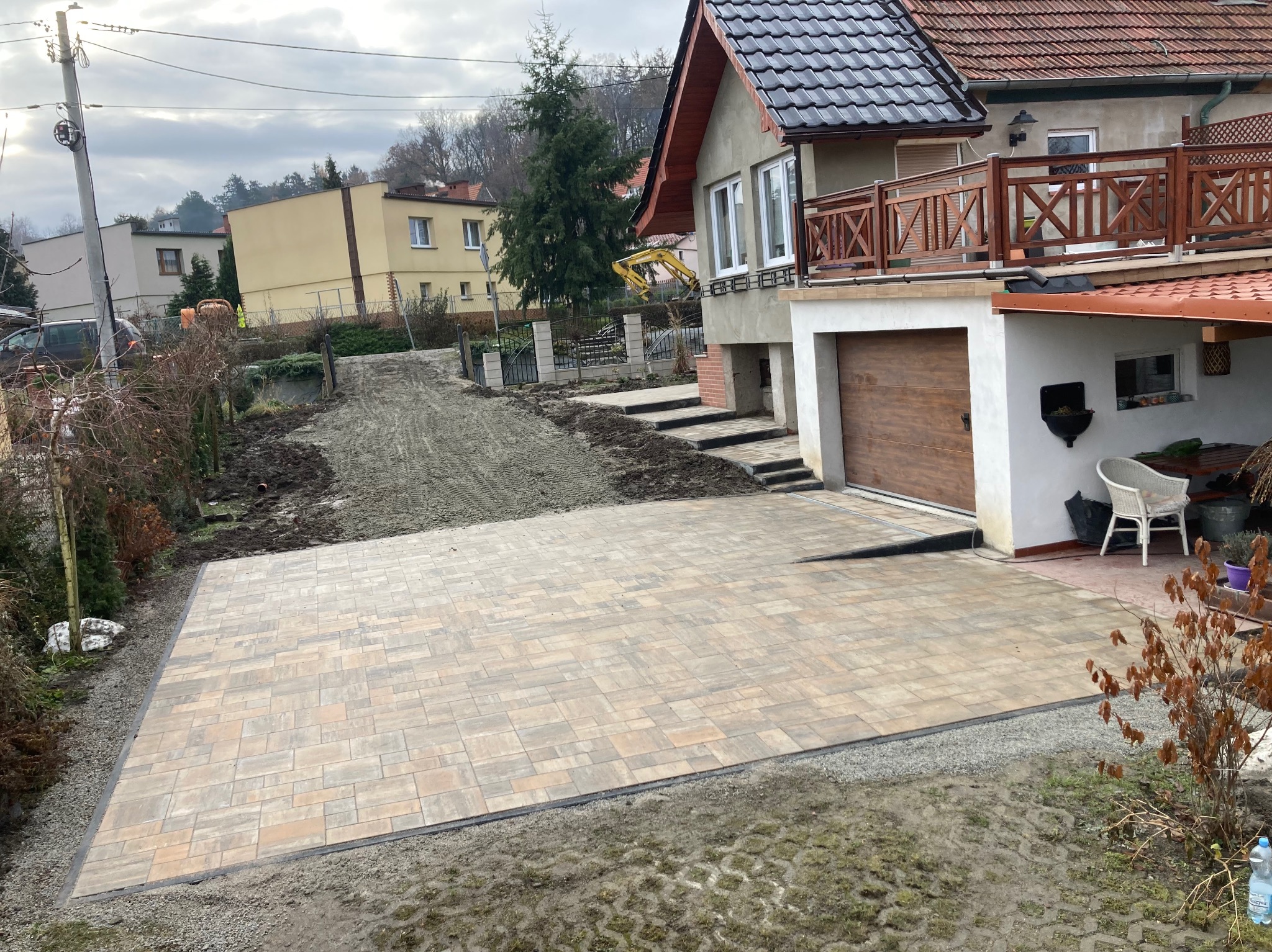 Ukończony podjazd z kostki brukowej w odcieniach beżu i brązu, z widocznymi obrzeżami, prowadzący do garażu domu jednorodzinnego; w tle przygotowany teren pod dalsze prace ziemne.