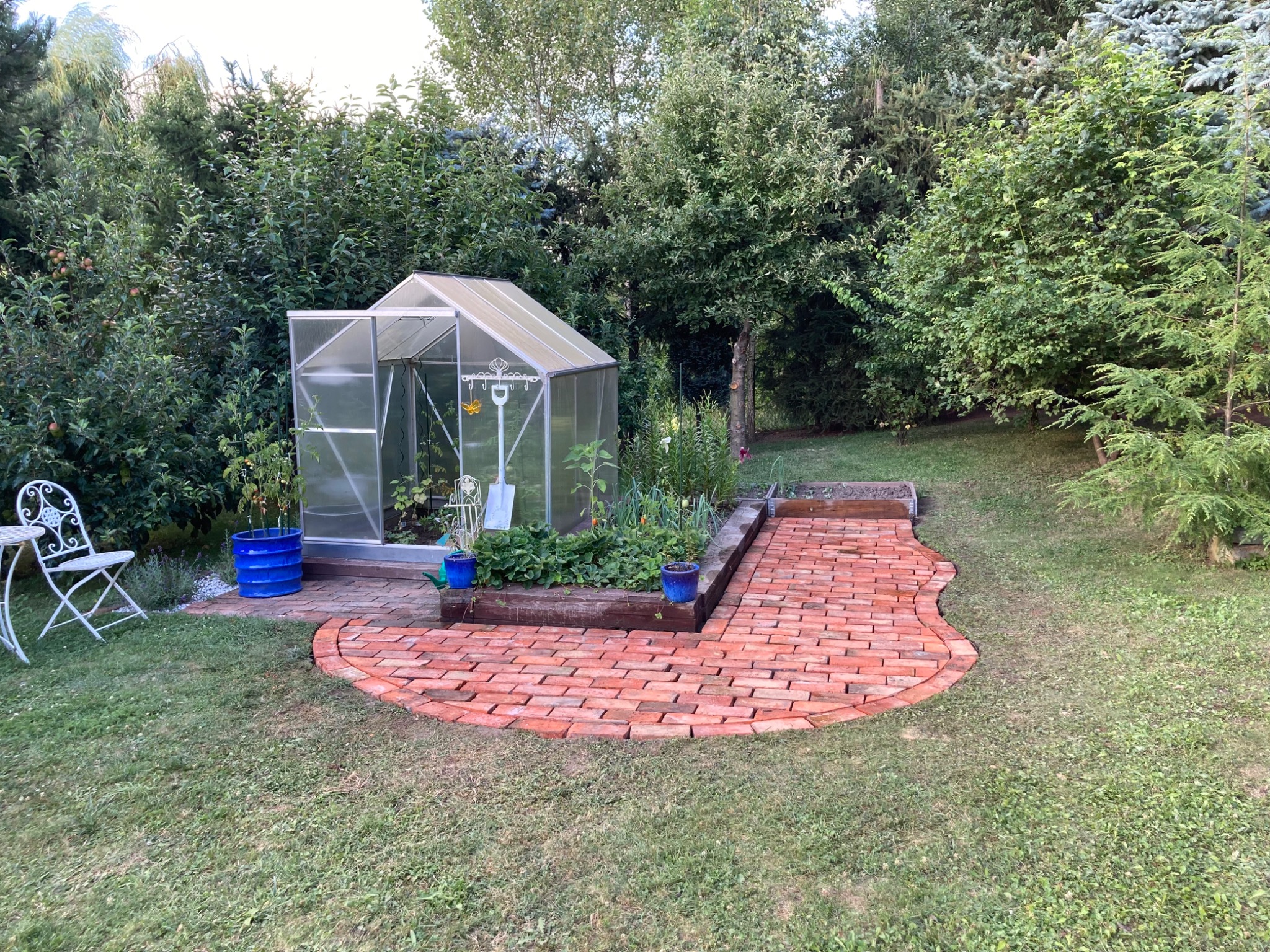 Ogród z ceglaną ścieżką prowadzącą do szklarni, otoczony zielenią drzew i krzewów, z metalowym stolikiem i krzesłem w tle.
