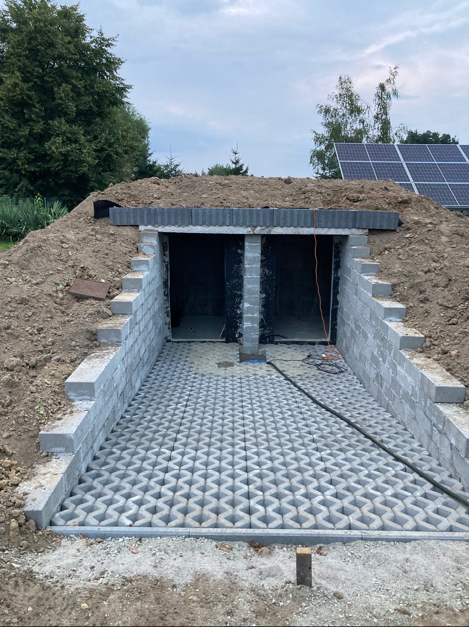 Wykop ziemny z dwoma wejściami wzmocnionymi betonowymi bloczkami, przed wejściami ułożona ażurowa nawierzchnia z betonowych płyt, w tle panele słoneczne.