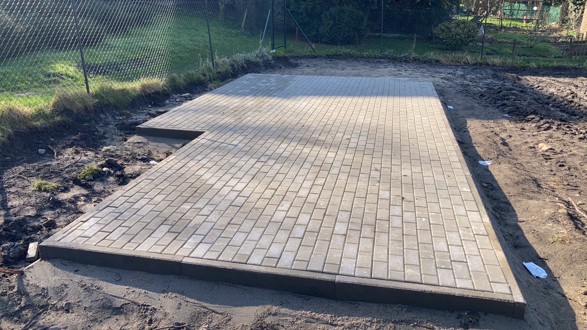 Ułożona, prostokątna powierzchnia z szarej kostki brukowej na przygotowanym podłożu, otoczona ziemią i ogrodzeniem z siatki.