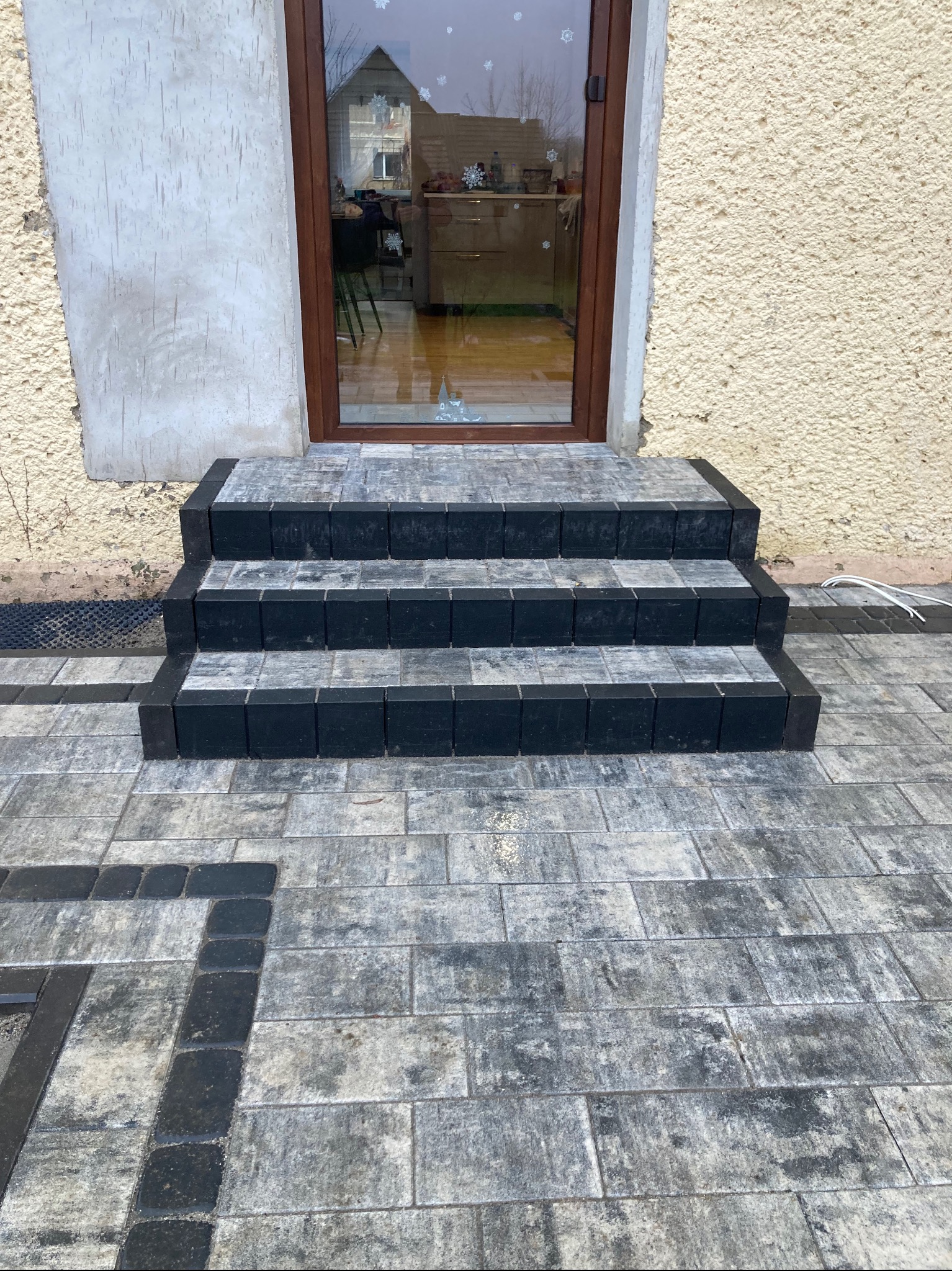 Schody wejściowe z kostki brukowej w odcieniach szarości i czerni, prowadzące do brązowych drzwi, z widocznym fragmentem elewacji o nierównej fakturze.