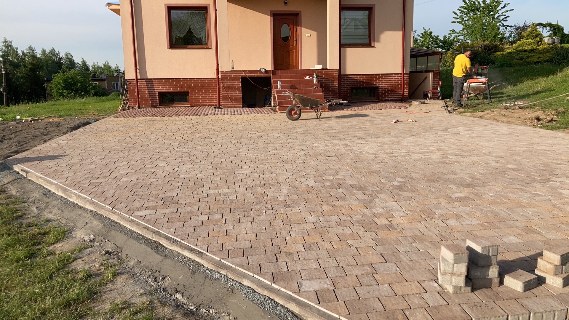 Ułożona kostka brukowa w kolorze beżowo-brązowym przed domem jednorodzinnym; na pierwszym planie stos kostki, w tle pracownik przy maszynie do cięcia kostki.