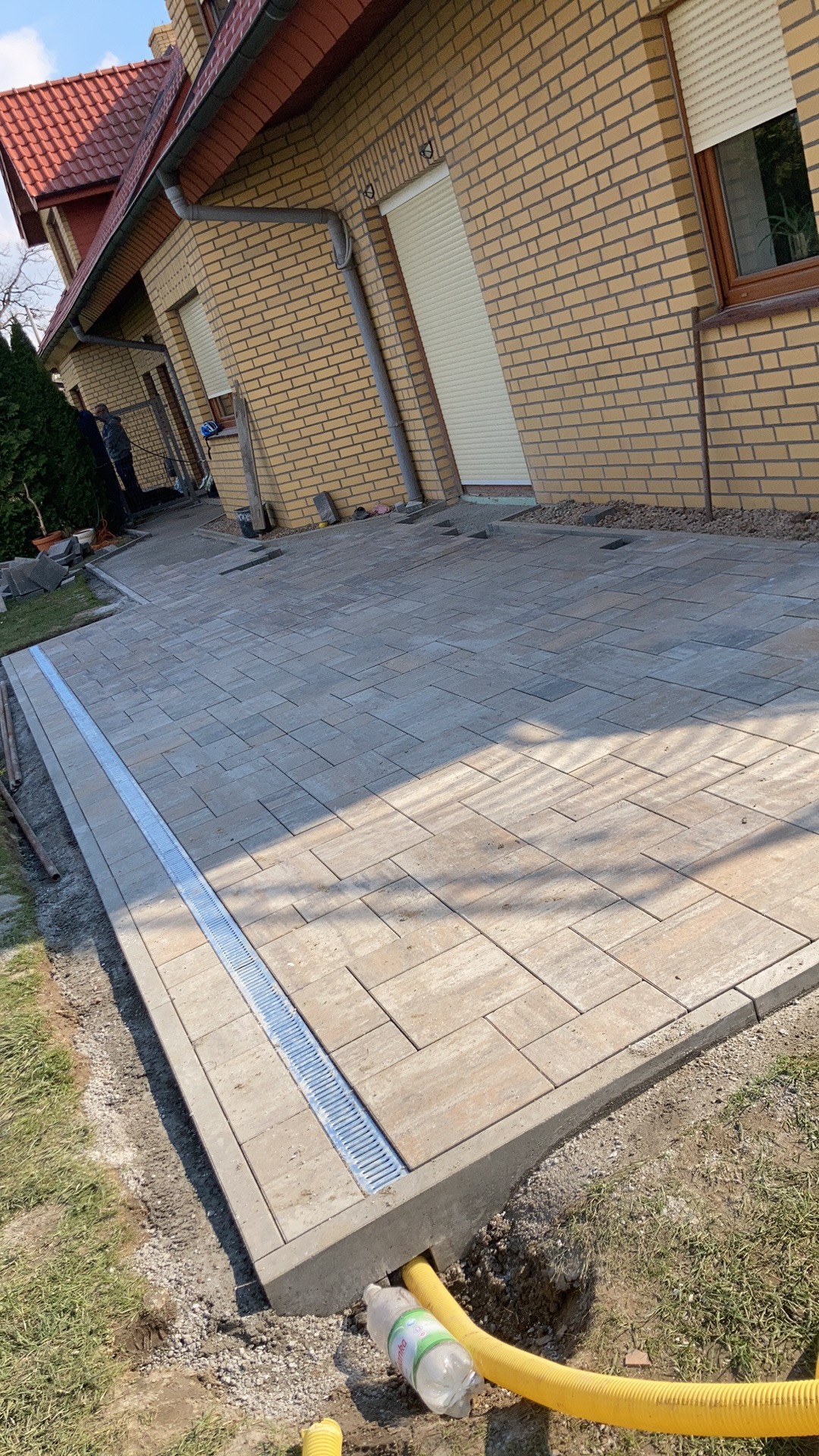 Fragment nowej nawierzchni z kostki brukowej, z widocznym odpływem liniowym i żółtą rurą drenażową, częściowo zakrytą butelką po wodzie.