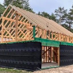 Konstrukcja drewniana domu w trakcie budowy, widoczna więźba dachowa i szkielet ścian, zabezpieczone czarną folią. W tle las.