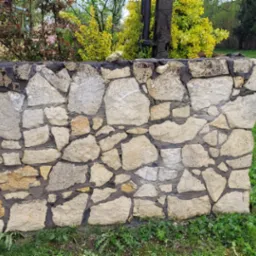 Kamienny murek z czarną skrzynką na listy, widoczny na tle zielonego trawnika, drzew i domu z jasną elewacją; obok murku stoi betonowy słup z numerem 21.