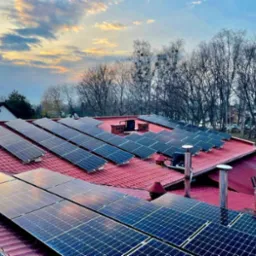 Panele słoneczne na czerwonym dachu domu jednorodzinnego, widok z góry, w tle zachodzące słońce i drzewa bez liści.