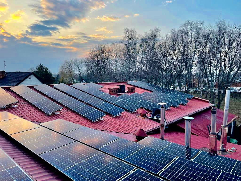 Panele słoneczne na czerwonym dachu domu jednorodzinnego, widok z góry, w tle zachodzące słońce i drzewa bez liści.