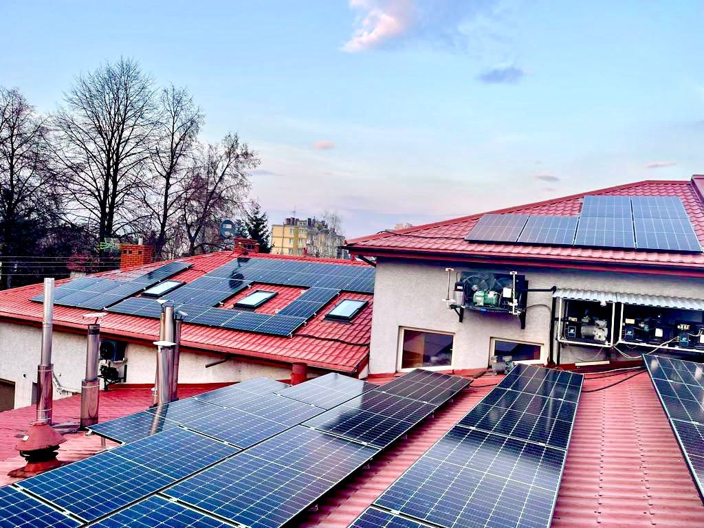 Panele słoneczne na czerwonym dachu domu z widocznymi oknami dachowymi i urządzeniami na ścianie, w tle budynki i drzewa.