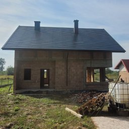 Usługi remontowo budowlane Andrzej Tapa - Surowy budynek jednorodzinny z ciemnoszarą dachówką ceramiczną i dwoma kominami, w trakcie budowy, widoczne niezabezpieczone otwory okienne i drzwiowe, paleta i zbiornik na wodę w tle.