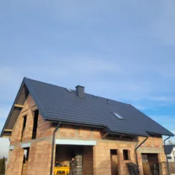 Surowy budynek jednorodzinny z ciemną dachówką ceramiczną, widoczne rynny i komin, brak okien, w tle błękitne niebo, dwóch robotników wewnątrz budynku przy żółtym stole roboczym.