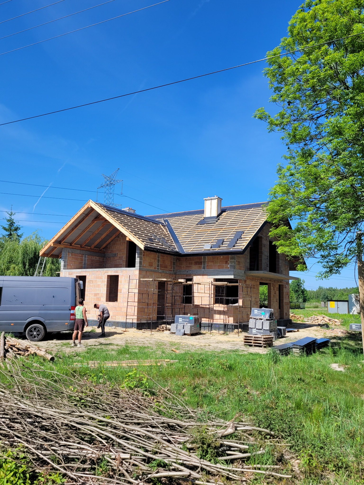 Budowa domu jednorodzinnego w trakcie, widoczna konstrukcja dachu z częściowo ułożoną dachówką, murarz i pomocnik obok szarego busa, palety z materiałami budowlanymi na trawniku.