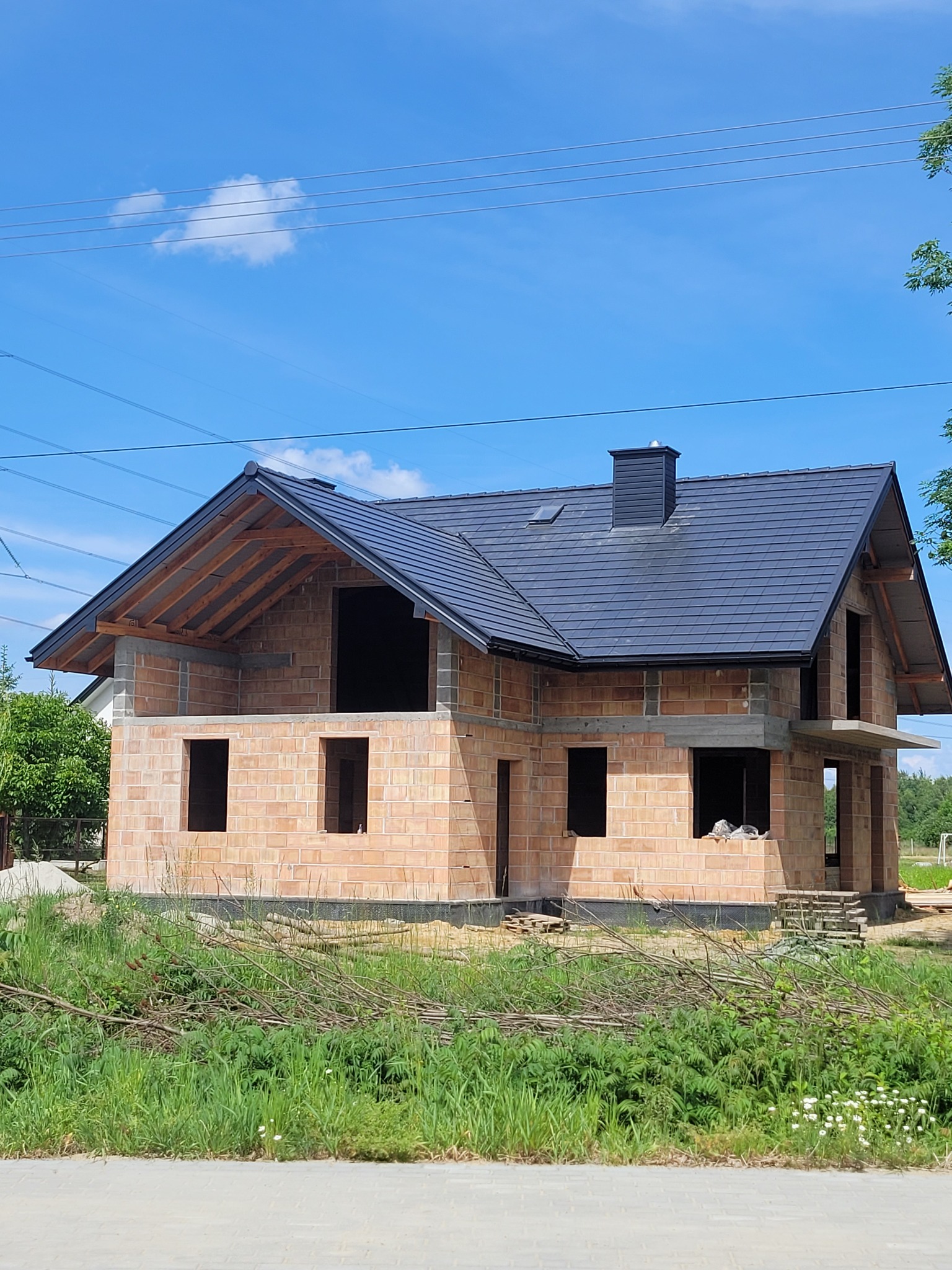 Dom w trakcie budowy z zamontowanym, grafitowym dachem z dachówki ceramicznej i kominem, widoczne drewniane elementy więźby dachowej, ściany z czerwonej cegły i niezagospodarowany teren wokół.