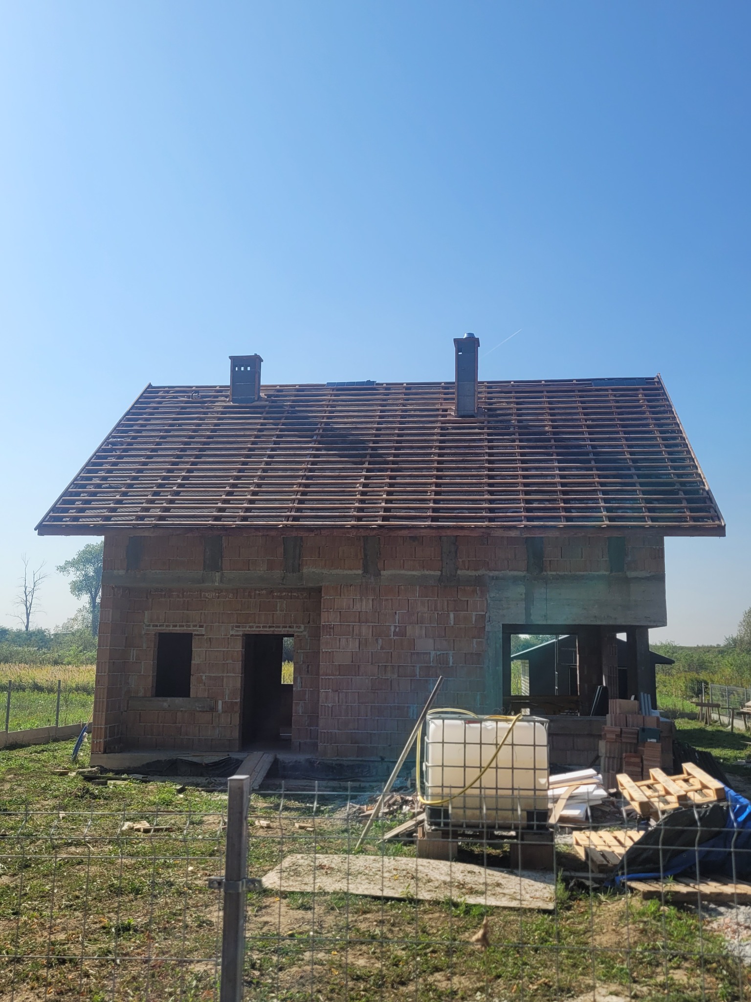 Niedokończony budynek z częściowo ułożoną więźbą dachową i kominem, widoczny zbiornik na wodę oraz materiały budowlane na posesji.