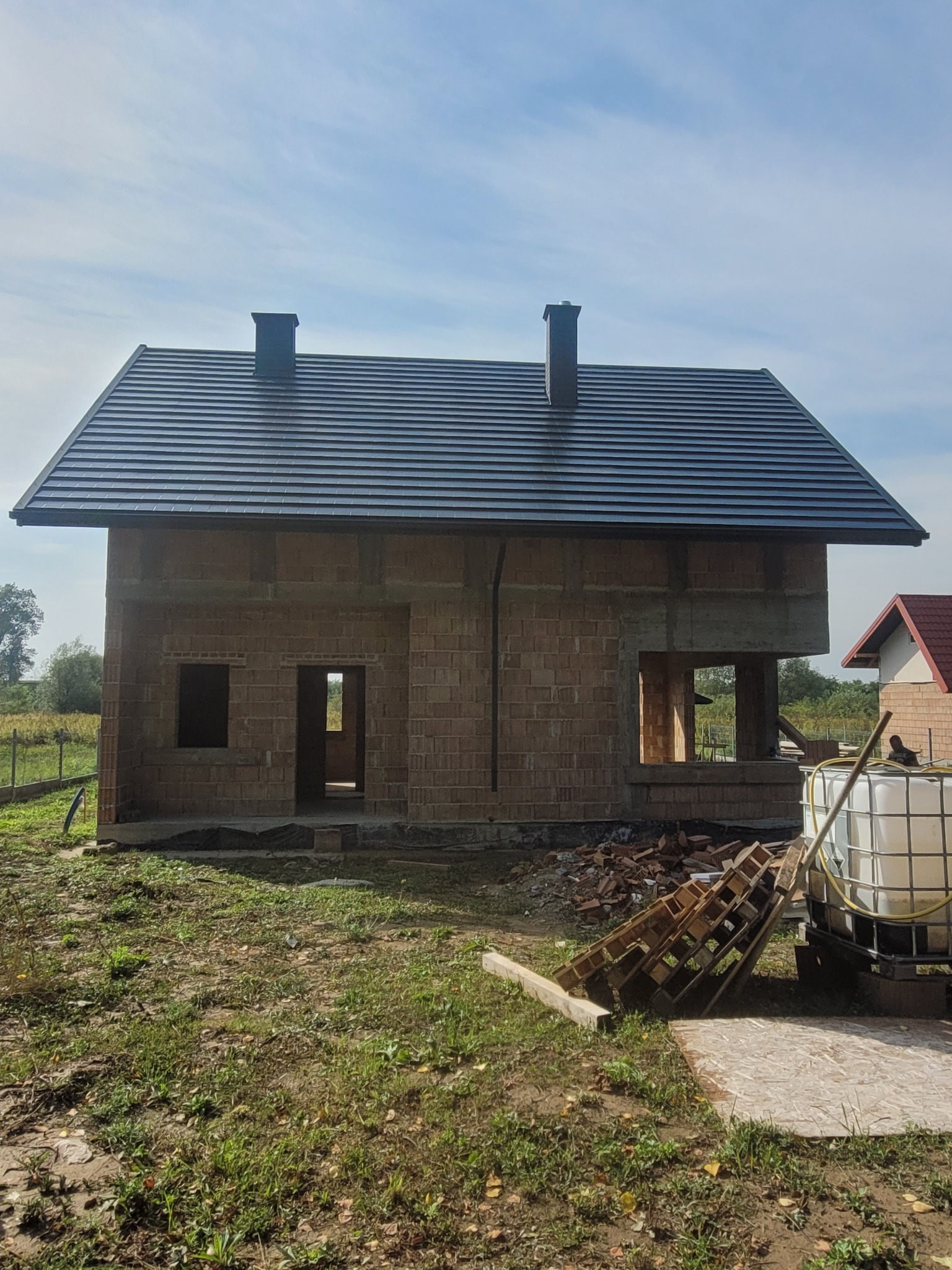 Surowy budynek jednorodzinny z ciemnoszarą dachówką ceramiczną i dwoma kominami, w trakcie budowy, widoczne niezabezpieczone otwory okienne i drzwiowe, paleta i zbiornik na wodę w tle.