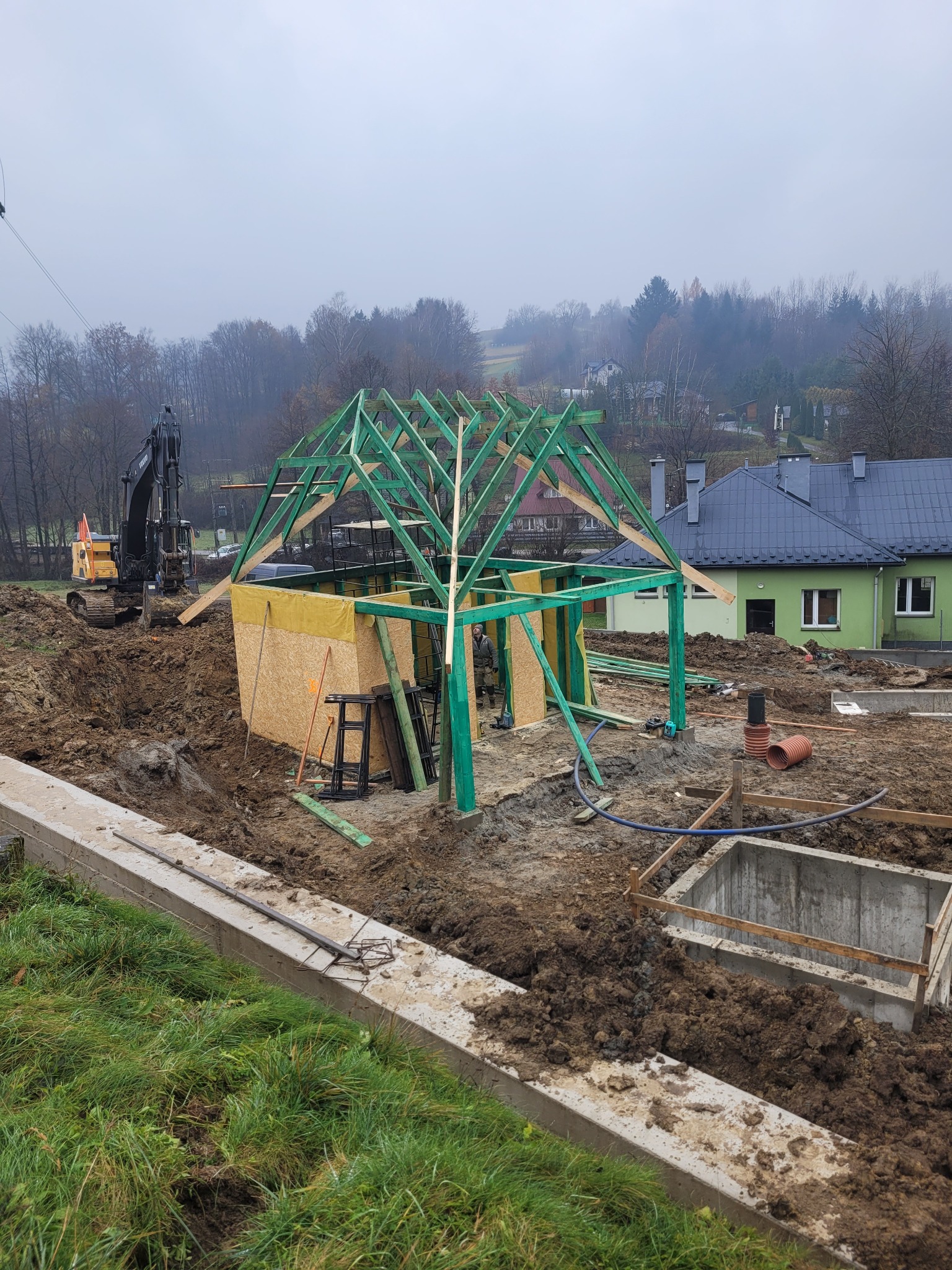 Budowa domu w trakcie wznoszenia konstrukcji szkieletowej dachu, widoczny szkielet z zielonych belek i żółta membrana, w tle koparka i zabudowania.