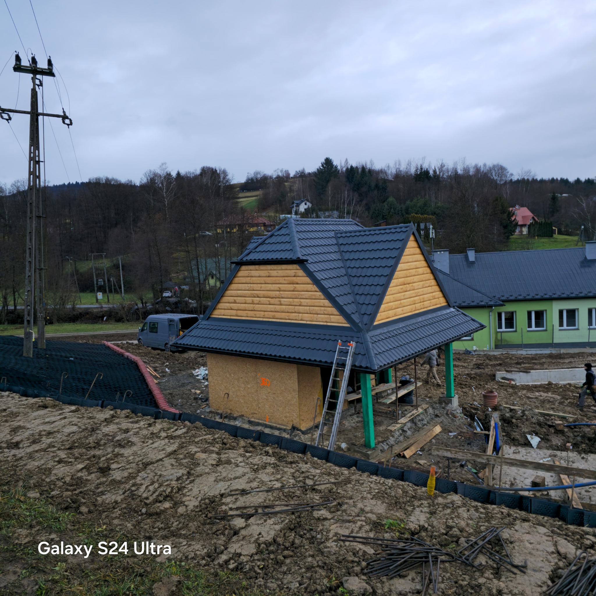 Konstrukcja altany z drewnianą elewacją i grafitowym pokryciem dachowym w trakcie budowy, widoczna drabina oparta o dach, w tle budynek i krajobraz.