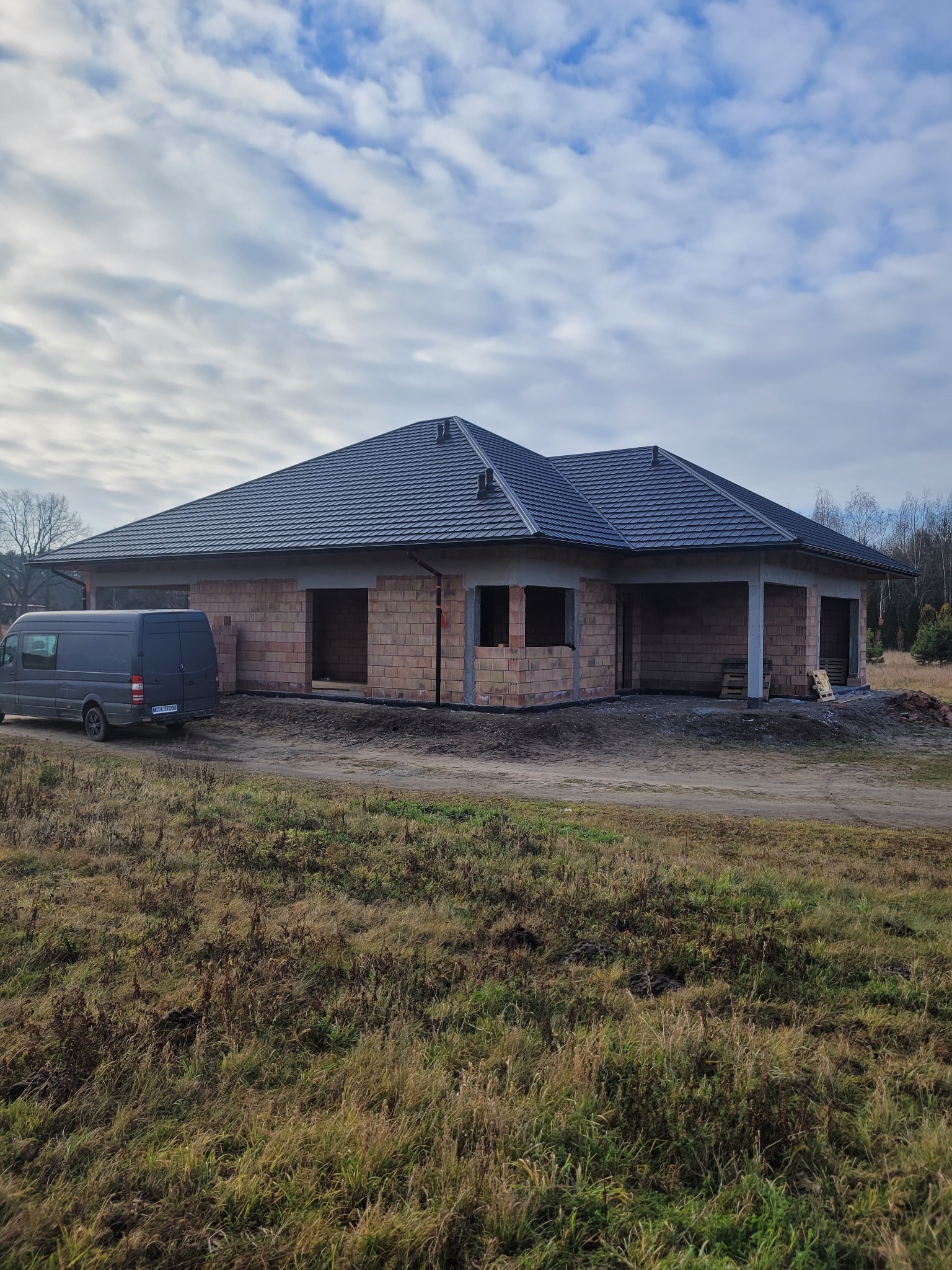 Niedokończony dom jednorodzinny z ciemną dachówką, widoczny szary bus na tle budynku w stanie surowym otwartym.