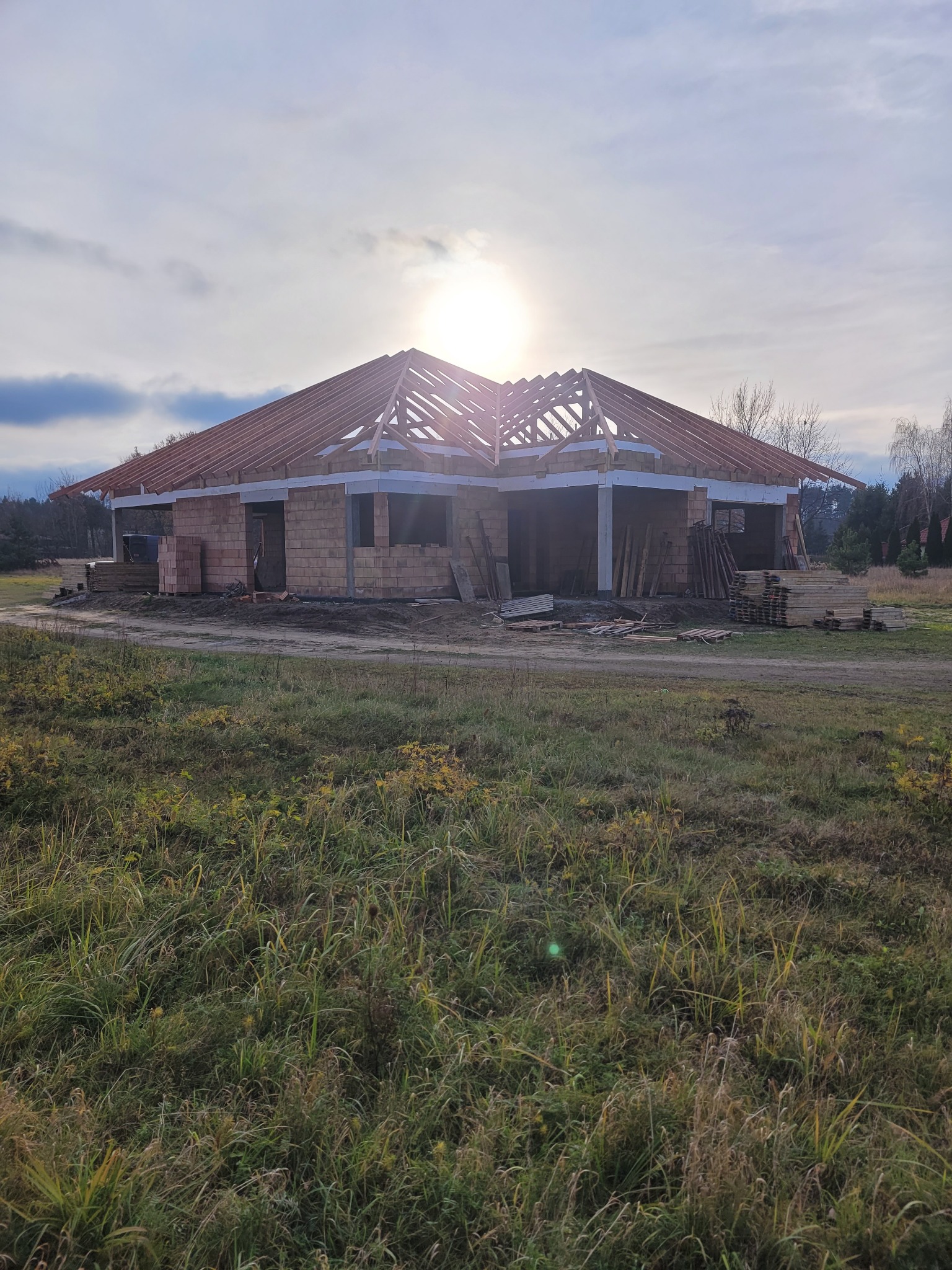 Dom jednorodzinny w trakcie budowy, widoczna konstrukcja drewniana dachu z częściowo ułożoną dachówką, ściany z bloczków, otoczenie polne.