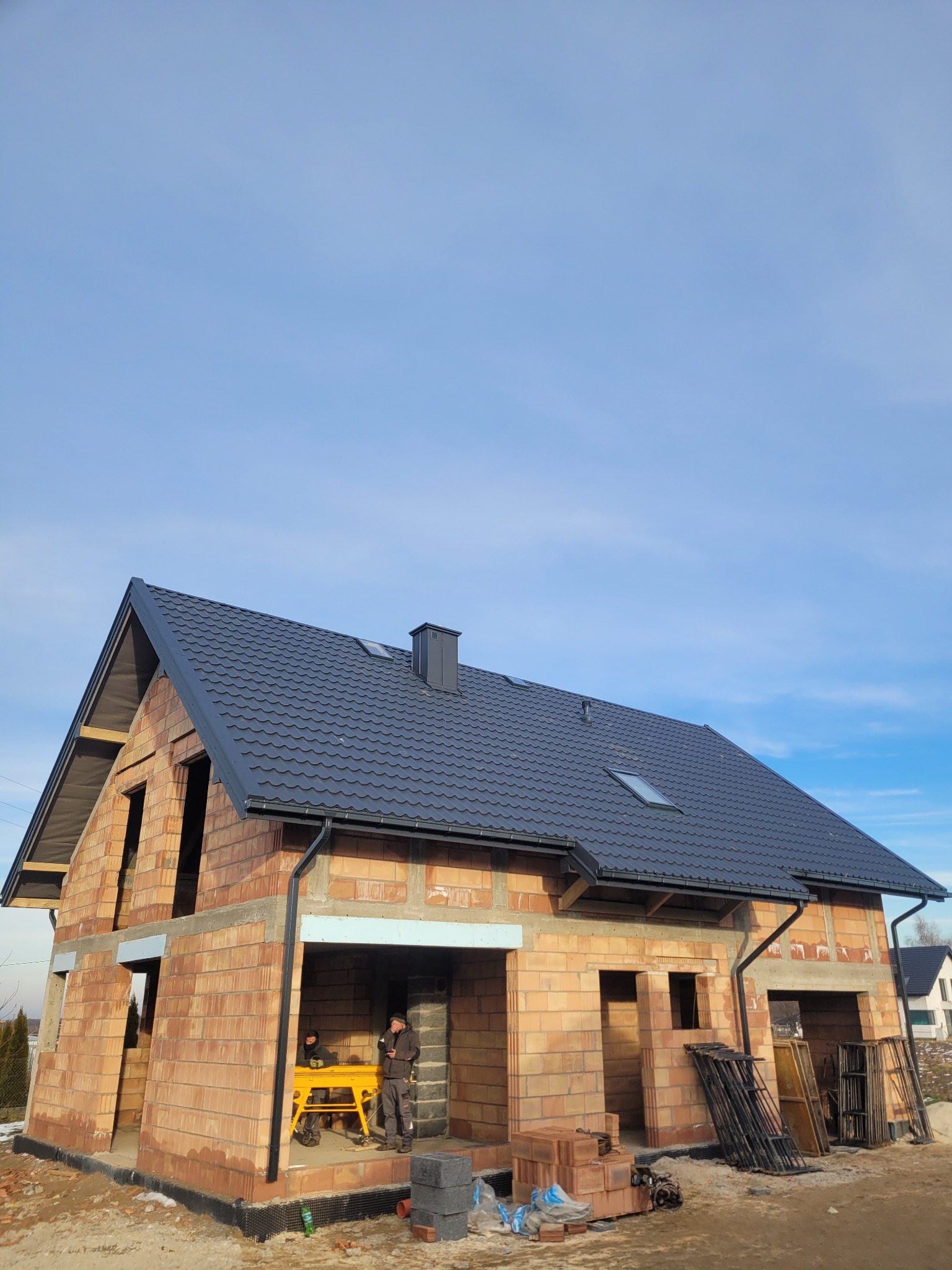 Surowy budynek jednorodzinny z ciemną dachówką ceramiczną, widoczne rynny i komin, brak okien, w tle błękitne niebo, dwóch robotników wewnątrz budynku przy żółtym stole roboczym.