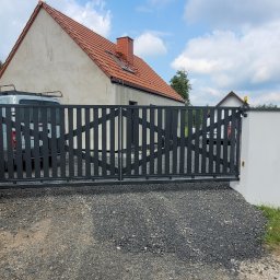 Karol Górny usługi spawalnicze - Bramy - Balustrady - Nowoczesna, ciemnoszara brama wjazdowa dwuskrzydłowa z pionowymi lamelami i ukośnymi elementami, osadzona w białych murkach, na tle domu z czerwonym dachem.