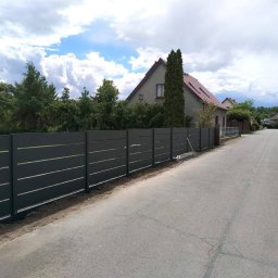 Karol Górny usługi spawalnicze - Bramy - Balustrady - Nowoczesne, ciemnoszare ogrodzenie panelowe z poziomymi lamelami, świeżo zamontowane wzdłuż asfaltowej drogi, z domem i zielenią w tle.