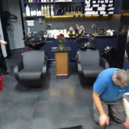 Wnętrze salonu fryzjerskiego z dwoma stanowiskami do mycia włosów, mężczyzna w niebieskiej koszulce czyści fotel, w tle kobieta z blond włosami i czerwone wiadro.