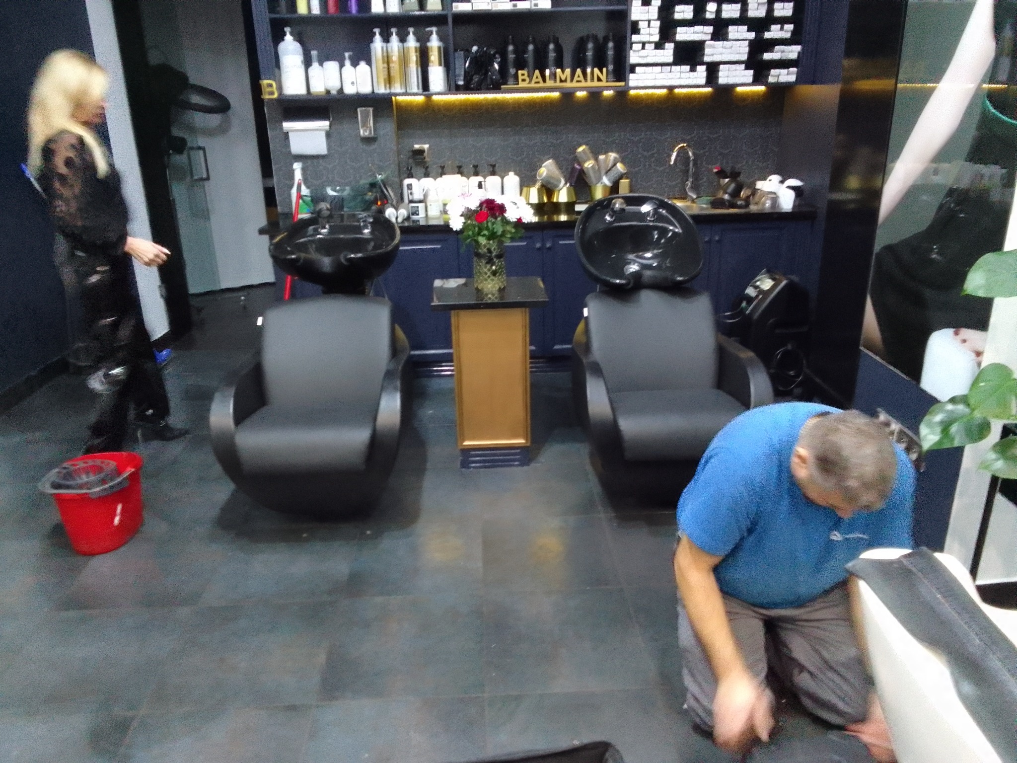 Wnętrze salonu fryzjerskiego z dwoma stanowiskami do mycia włosów, mężczyzna w niebieskiej koszulce czyści fotel, w tle kobieta z blond włosami i czerwone wiadro.