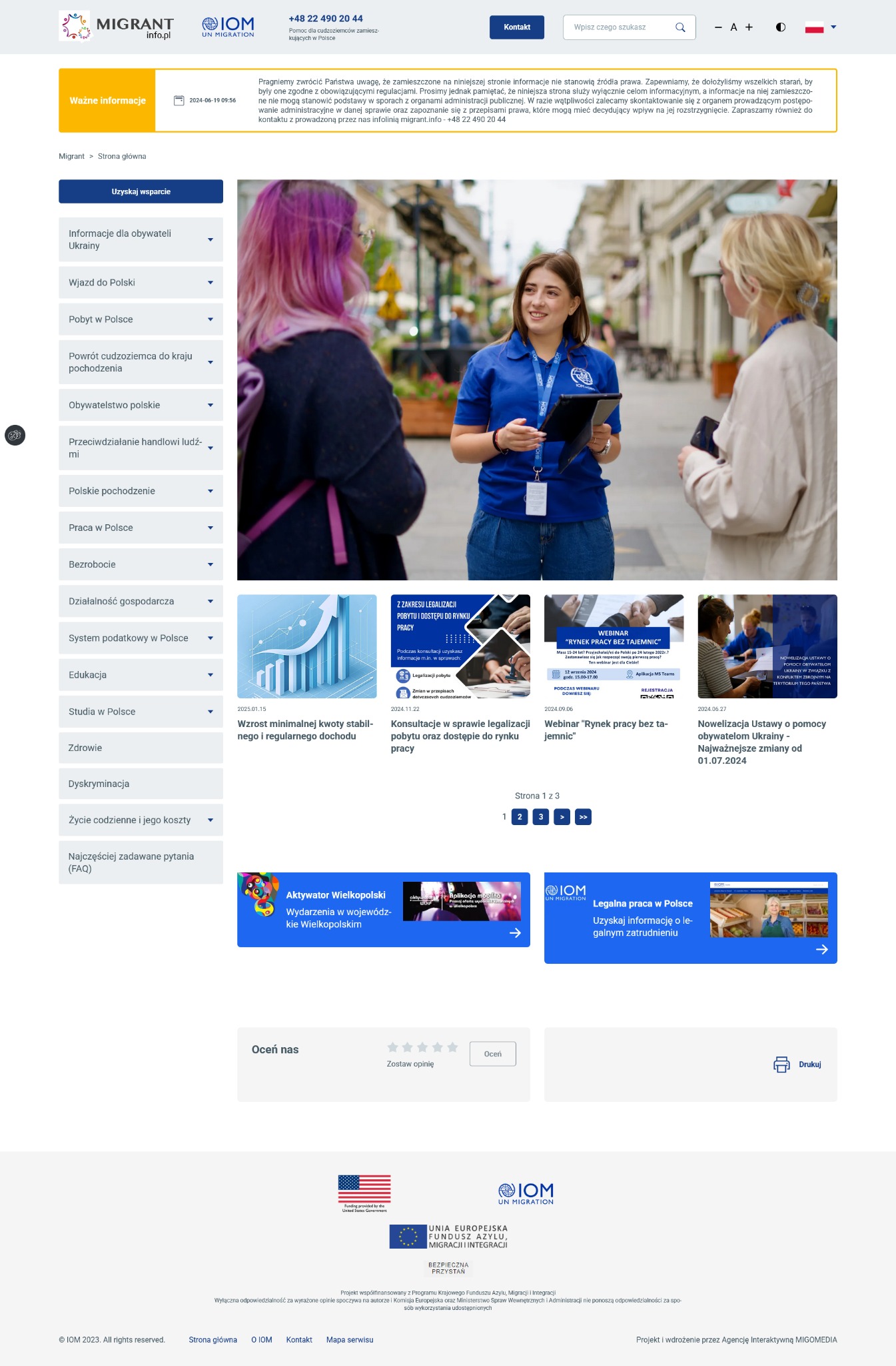 Dwie kobiety w przestrzeni miejskiej, jedna w niebieskiej koszulce z logo IOM, trzyma tablet, druga z różowymi włosami słucha, prawdopodobnie rozmawiają o sprawach związanych z migracją.